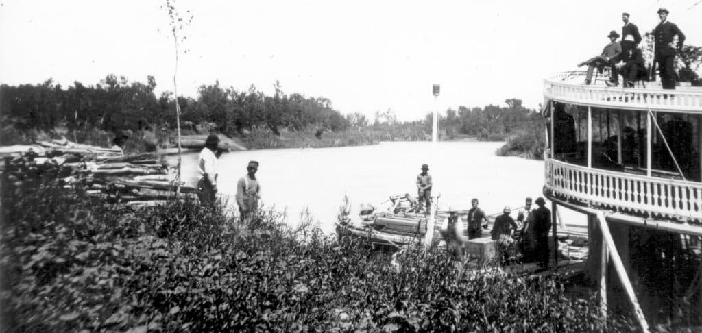 This is a black and white photo of men standing on a large boat. There are man standing on land, with lumber.