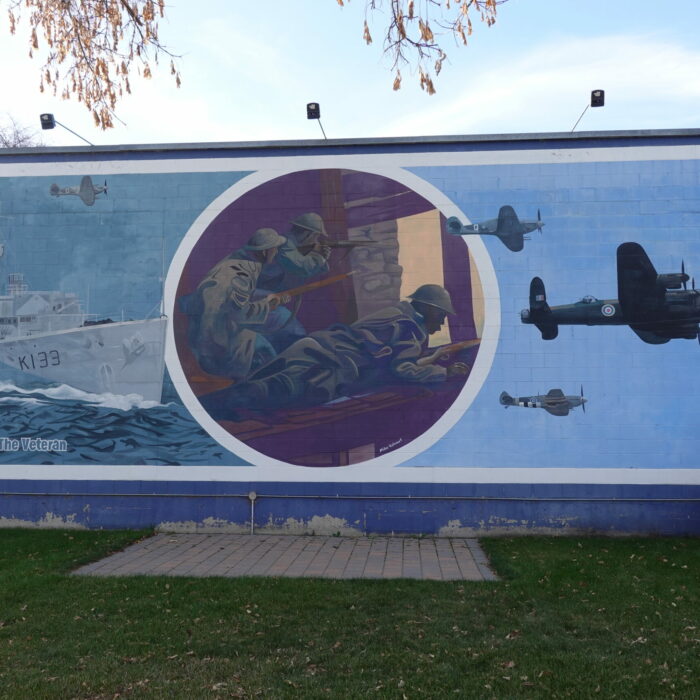 Colour photo of a large mural on the side of a building. The mural is of a naval ship on the left side, a few soldiers in the center circle, and airplanes on the right side