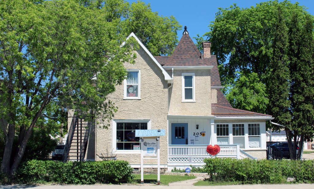 A colour image of a cottage style house with a turret on the right side above a porch. The house is beige and the porch and trim work is painted white