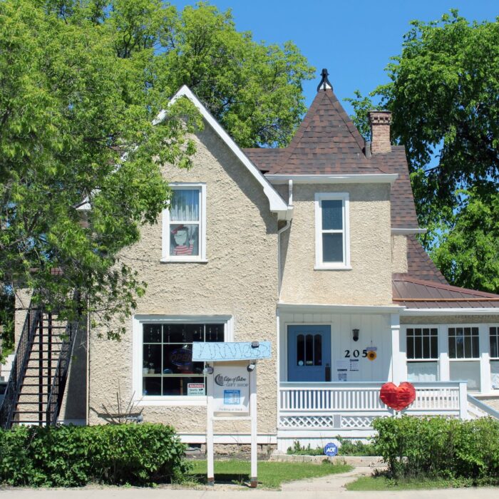 A colour image of a cottage style house with a turret on the right side above a porch. The house is beige and the porch and trim work is painted white