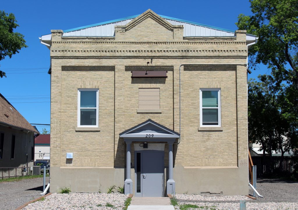 Colour image of a symmetrical brick building. The front door is set down a flight of stairs lower than street level. There are two windows on either side of the door and a peaked roof