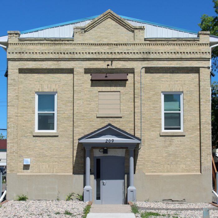 Colour image of a symmetrical brick building. The front door is set down a flight of stairs lower than street level. There are two windows on either side of the door and a peaked roof