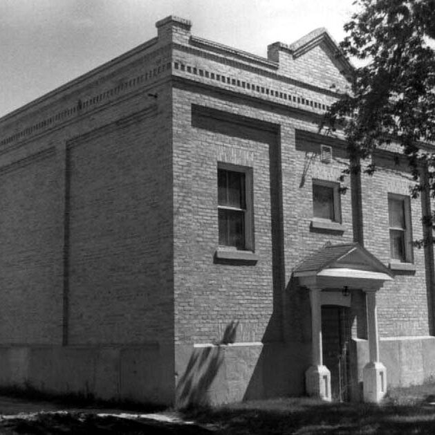 A black and white image of a symmetrical brick building with two windows beside the front door and a third above the front door.