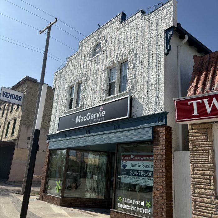 Colour photo of a store front. The ground floor of the building is brick with a blue horizontal trim piece between the storeys. The second storey stucco is painted white. There is brick trim around the windows