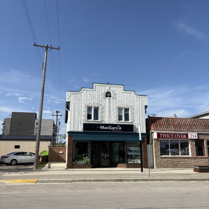 Colour photo of a store front. The ground floor of the building is brick with a blue horizontal trim piece between the storeys. The second story stucco is painted white. There is brick trim around the windows