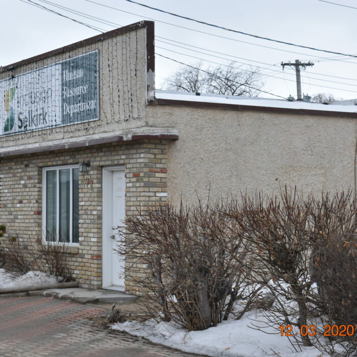 Colour photo of a small beige building with a tall facade. There is a white door and two windows on the front. There are lots of short trimmed shrubs on right side of the building