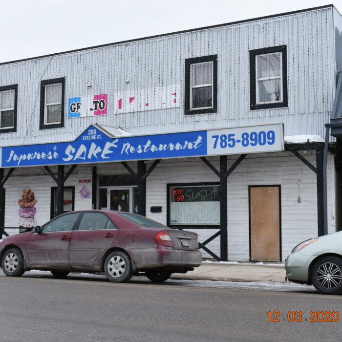 Colour photo of a two-story white building with a blue sign which reads Sake. There are cars parked along the front of the restaurant on the road