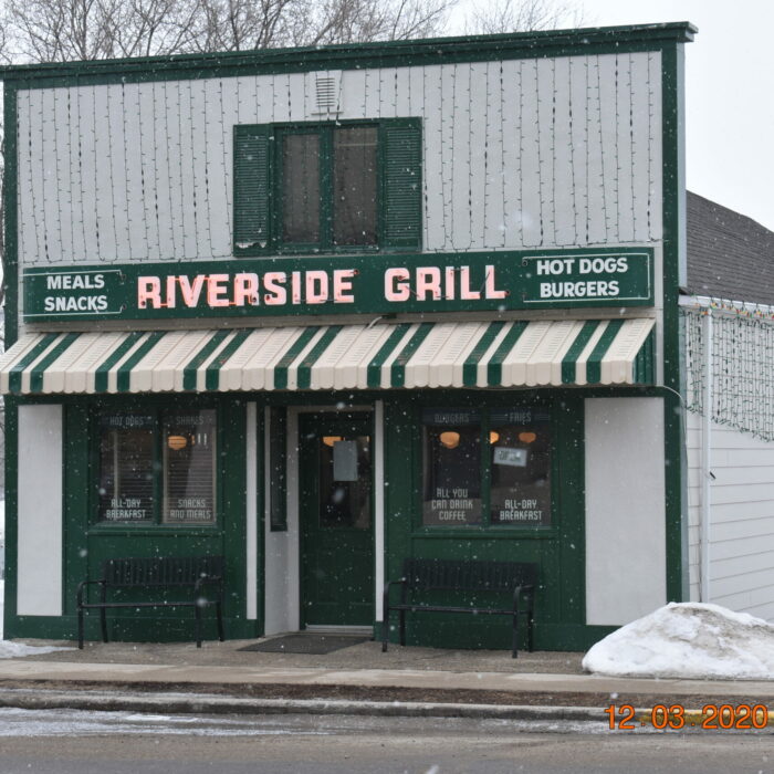 Colour photo of a small building with a boom-town style facade. The building is white with green trim. There is a stripped awning and a sign above the awning which reads 'Riverside Grill'