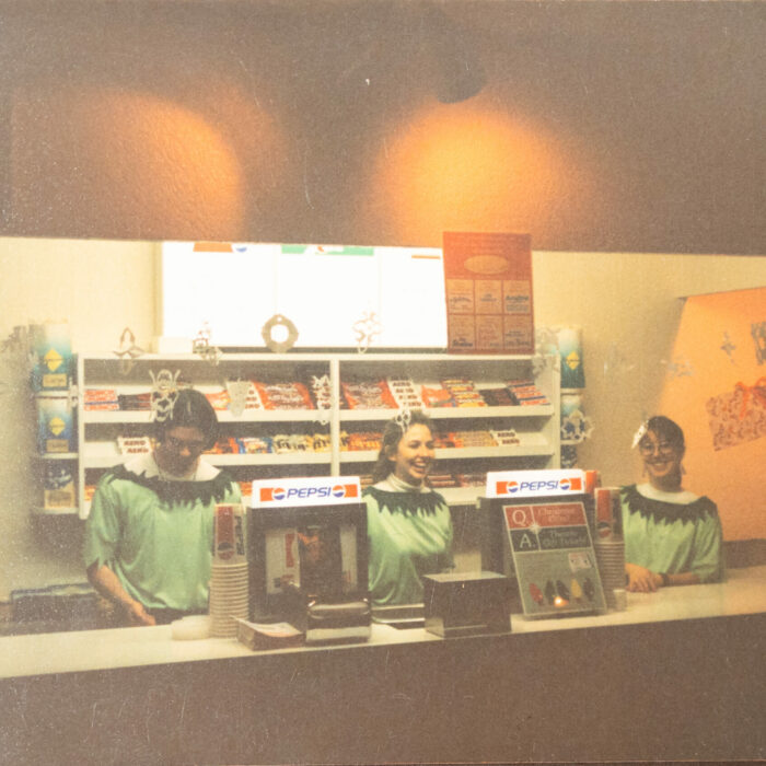 Colour photo of the canteen at the Garry Theatre. The lights are dim except for behind the counter where the lights are brighter. There are three people standing behind the counter