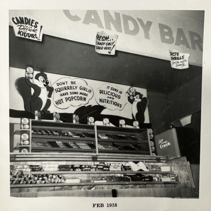 Black and white photo of a candy bar. There is a counter with a large display case. Behind the counter is a poster and a sign hung on the wall which says 'Candy Bar'