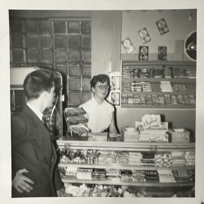 A black and white image of a young girl standing at a counter and display case filled with candies. A young man stands on the left hand side and looks at her