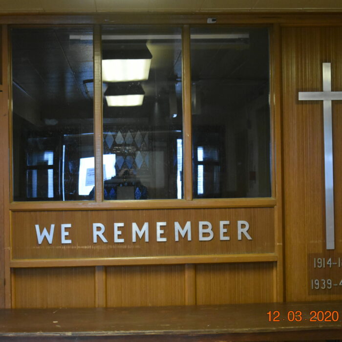 A colour photo of the back wall of the Memorial Hall Chapel. The words 'We Remember' are across the back below a set of three windows