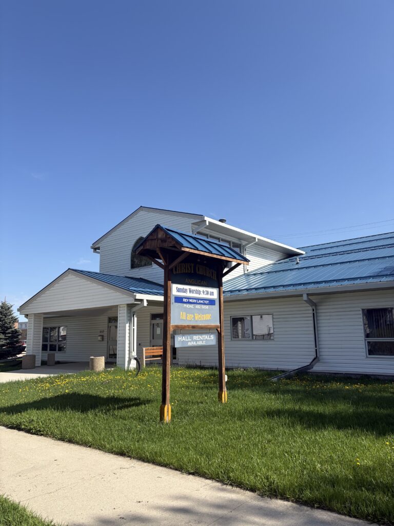 Colour photo of Christ Church exterior. The building is white with a blue tin roof. There is a sign at the front of the building