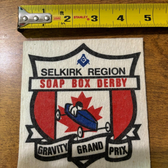 Colour image of a soap box derby crest. The crest has red on vertical panels on either side of a small blue derby car. Behind the derby car is a red canadian maple leaf. On top of the crest reads 'Selkirk Region Soap Box Derby' and at the bottom in a ribbon it reads 'gravity grand prix'