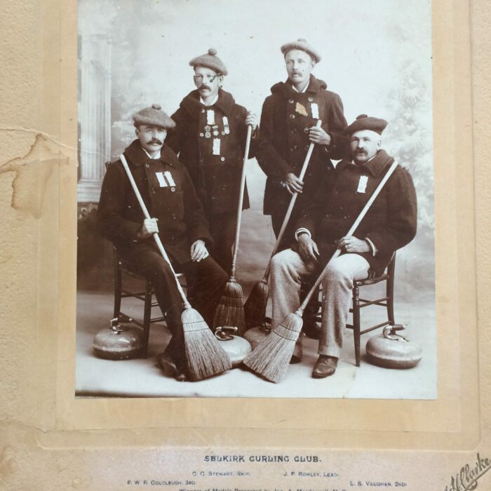 Black and white photo of four curling players. They hold brooms. Two sit and two stand. There are two curling rocks on the floor
