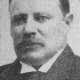 Black and white image of a man looking to the left of center. He is wearing a suit and has a moustache