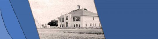 Eaton Headerbar This is a black and white photo of Devonshire School in 1919. it is a tall two story building with windows along each floor. in front of it is an empty field, and there are buildings to its left.