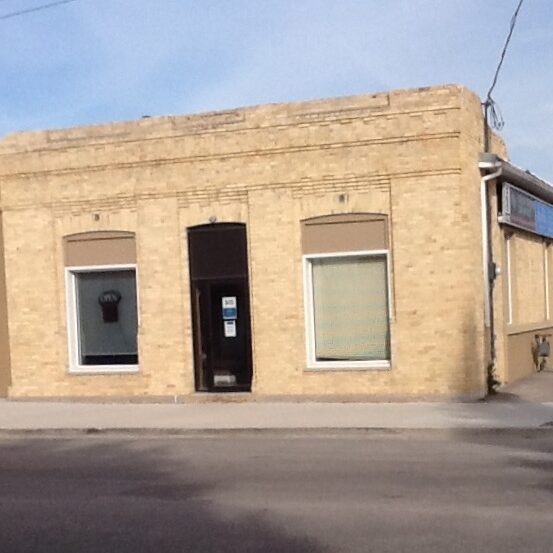 A colour image of a small brick building taken from across the street. The building is symmetrical with a two windows on either side of the front door
