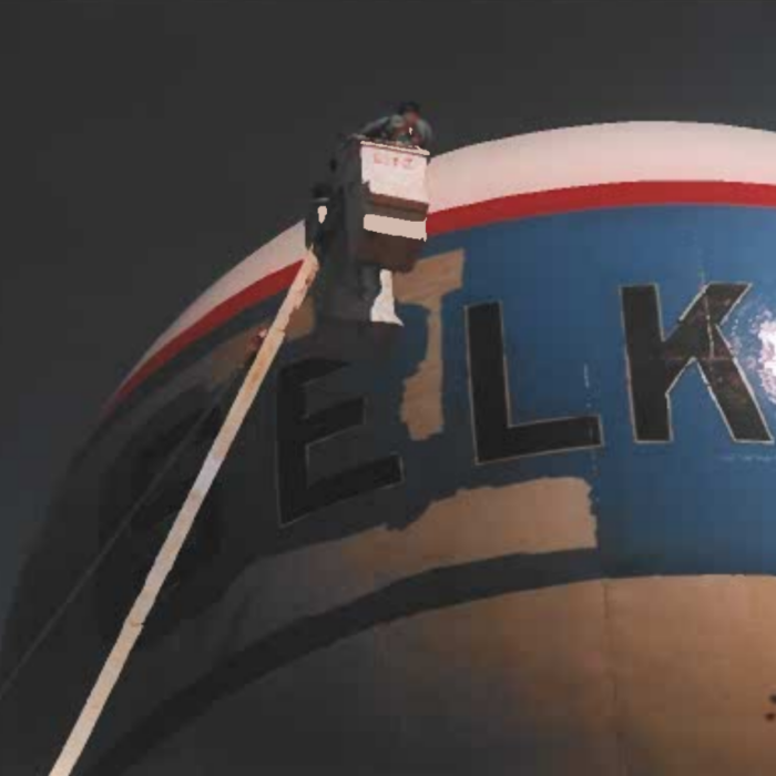 Colour photo of a crane raised up beside the water tower. The blue paint on the water tower is nearly completed.