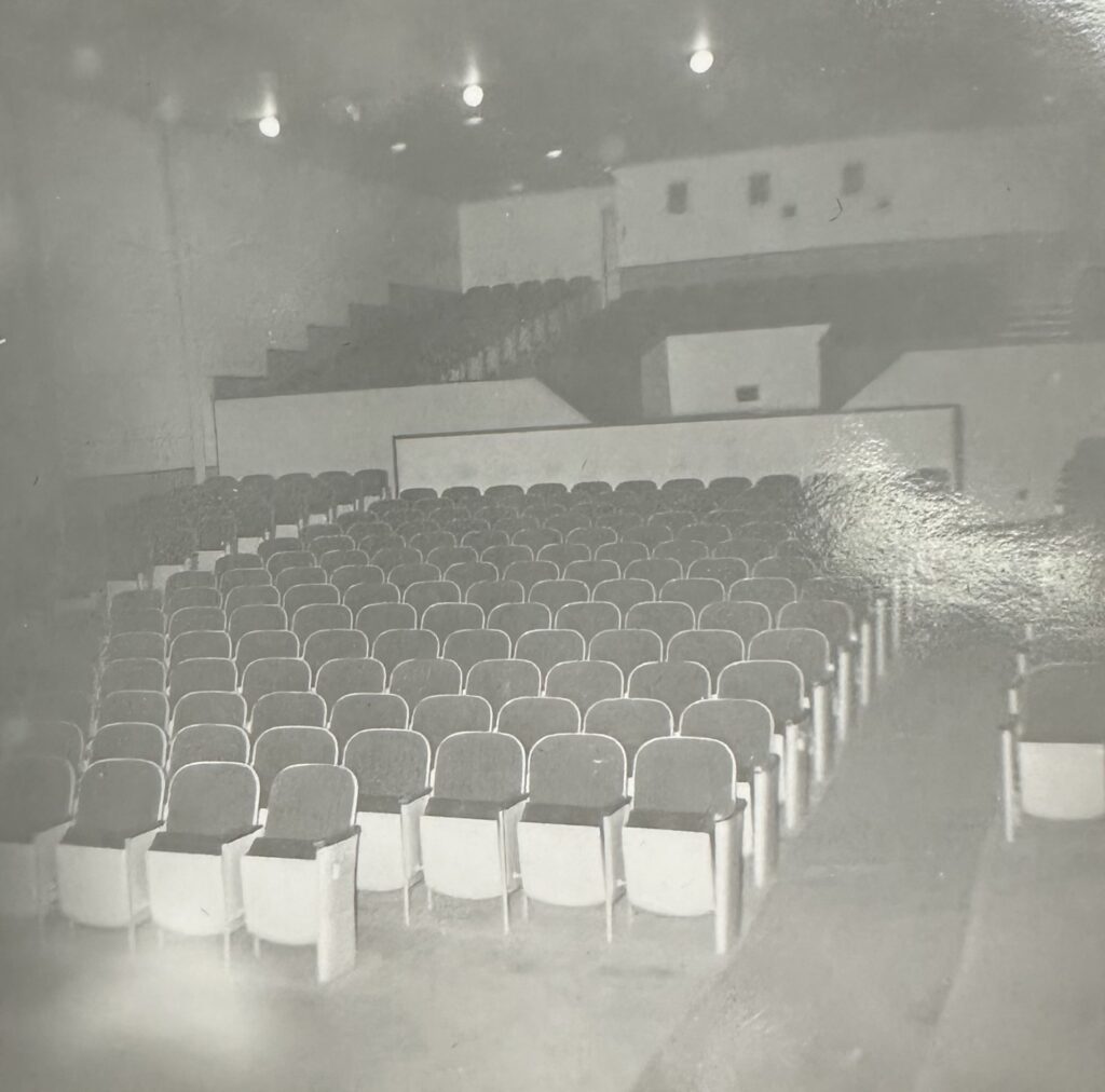 A black and white photo of a empty theatre. The seats rise up towards the top of the theatre. A few lights hang from the ceiling
