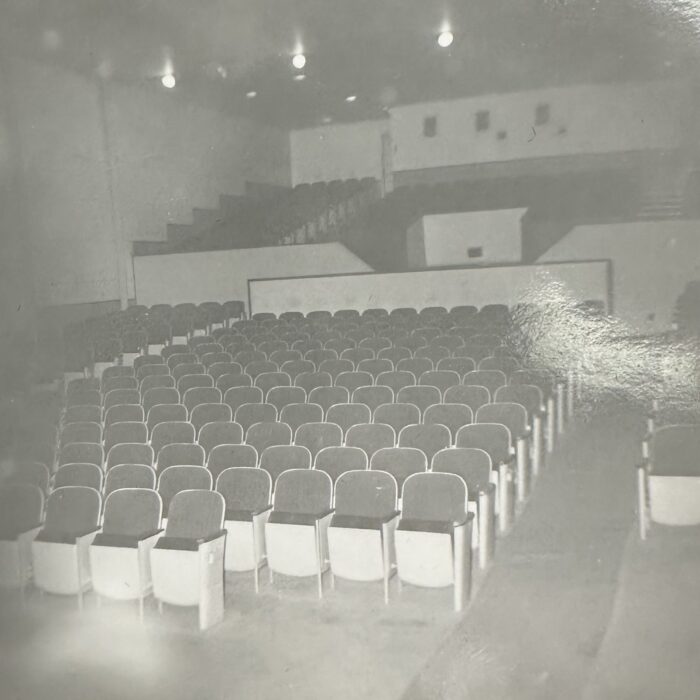A black and white photo of a empty theatre. The seats rise up towards the top of the theatre. A few lights hang from the ceiling