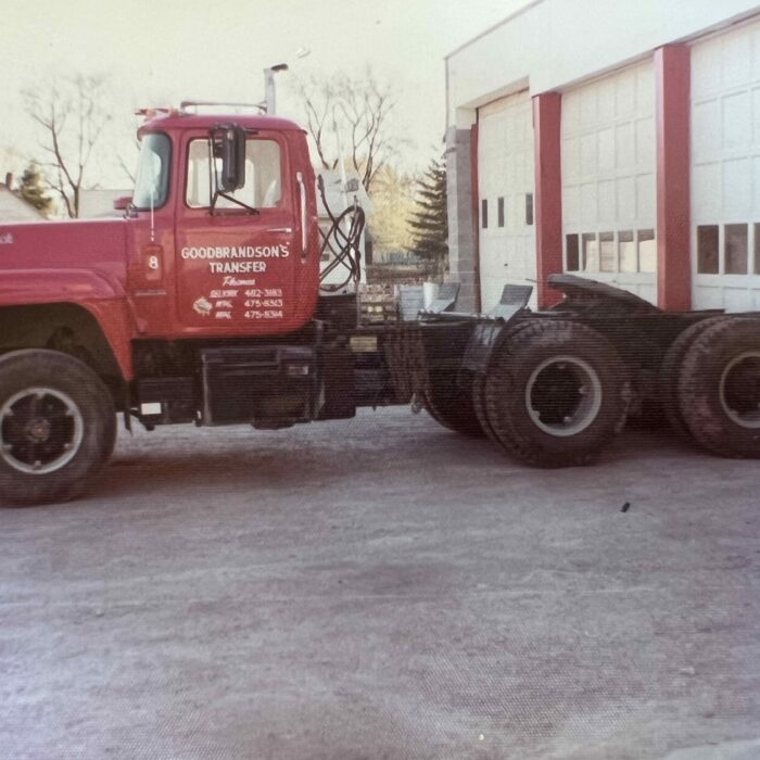 Colour photo of a red semi-truck parked in front of a white garage door.