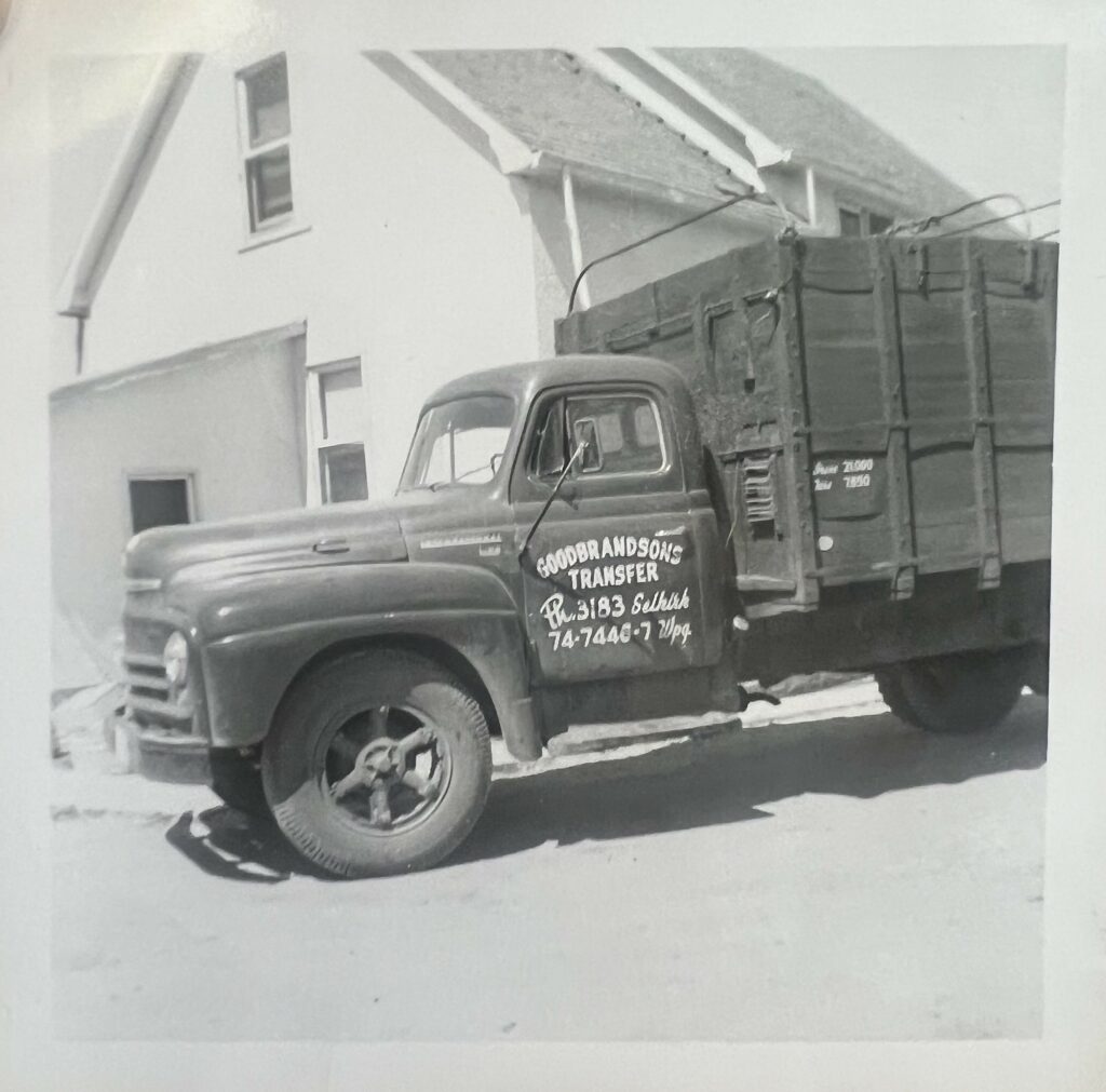 Black and white photo of a truck with a wooden truck box. There is a house in the background