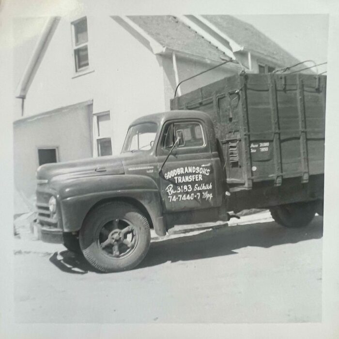Black and white photo of a truck with a wooden truck box. There is a house in the background