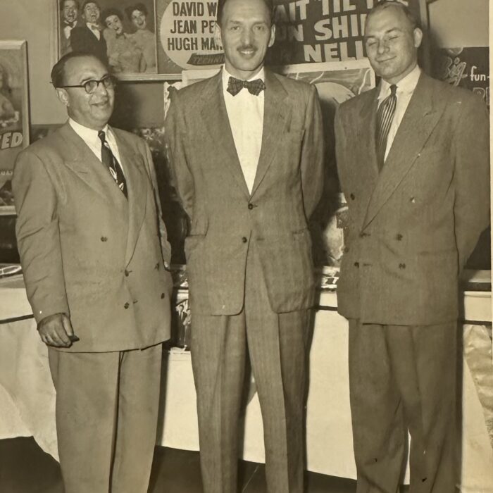 A black and white photo of three men standing in front of a counter. They are all wearing suits with ties and smile at the camera