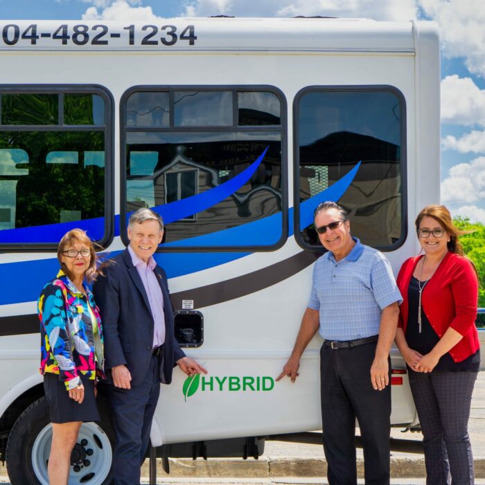 A colour photo of a the back half of a bus. In fron tof the bus are four people who point to the word 'hybrid' written in green on the bus