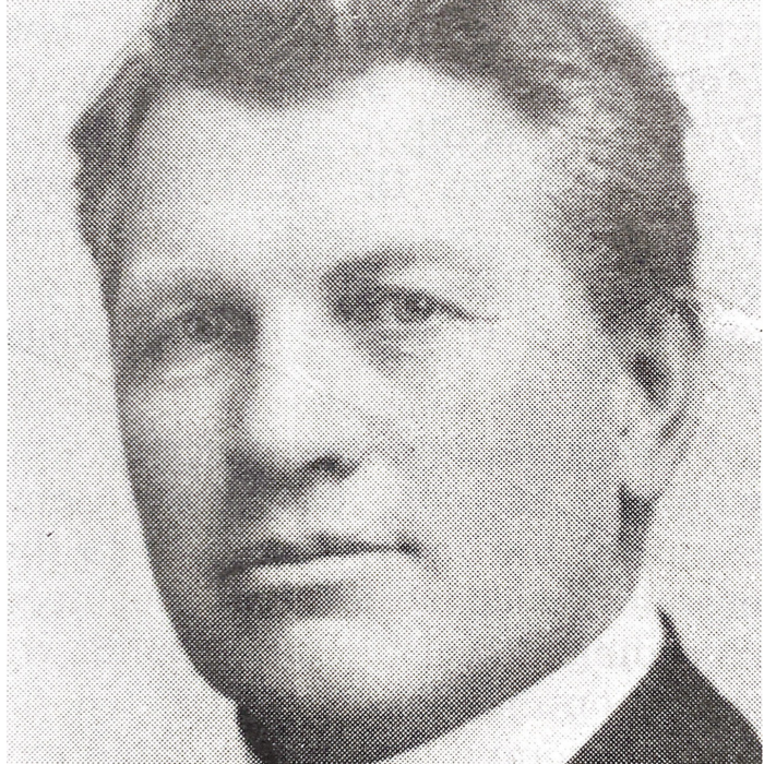 Black and white image of a man looking towards the camera. He is wearing a suit