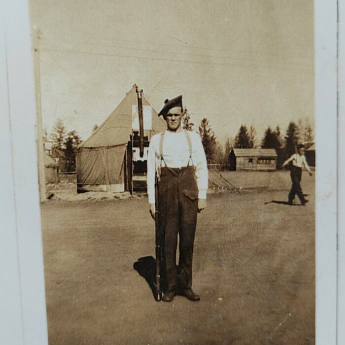 John V. Sinclair at Camp, c1942-1945, Mae Gulewich A sepia-coloured photo of a man standing in a grassy space. There is a tent in the background