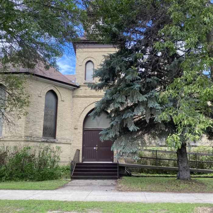 Brick building with tree and sidewalk in the foreground. Brick building has dark shingled roof with stairs leading to a arched door.
