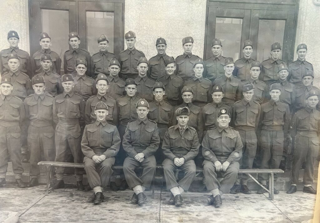 Black and white photo of a group of men dressed in military uniform. They are standing and sitting in front of a building