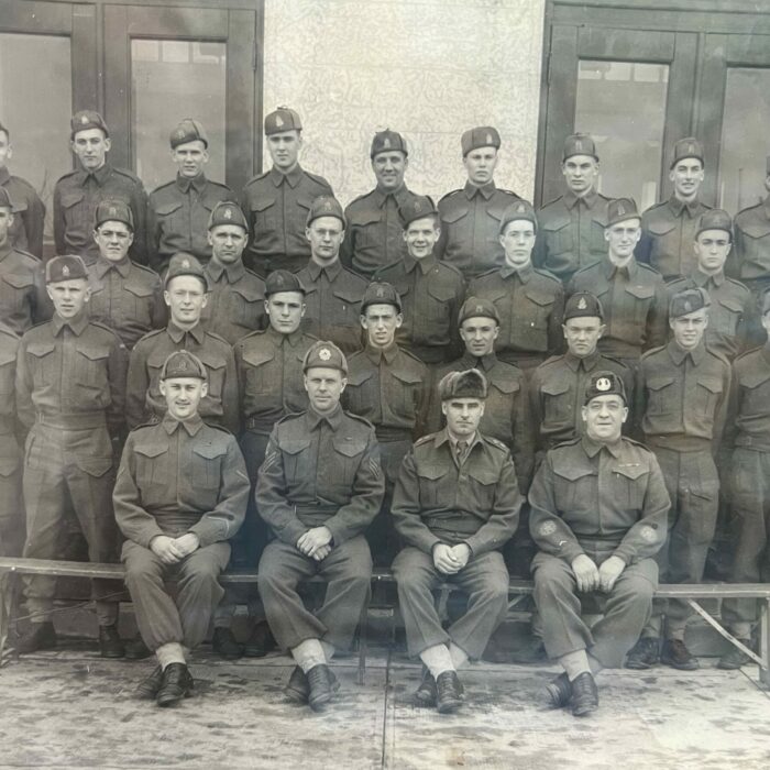 Black and white photo of a group of men dressed in military uniform. They are standing and sitting in front of a building