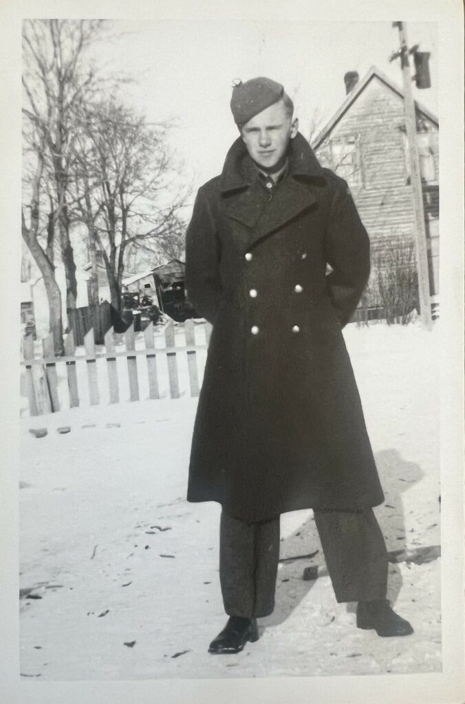 Black and white photo of Lefty standing in the snow. He is wearing a peacoat with double buttons down the front. He is also wearing a hat
