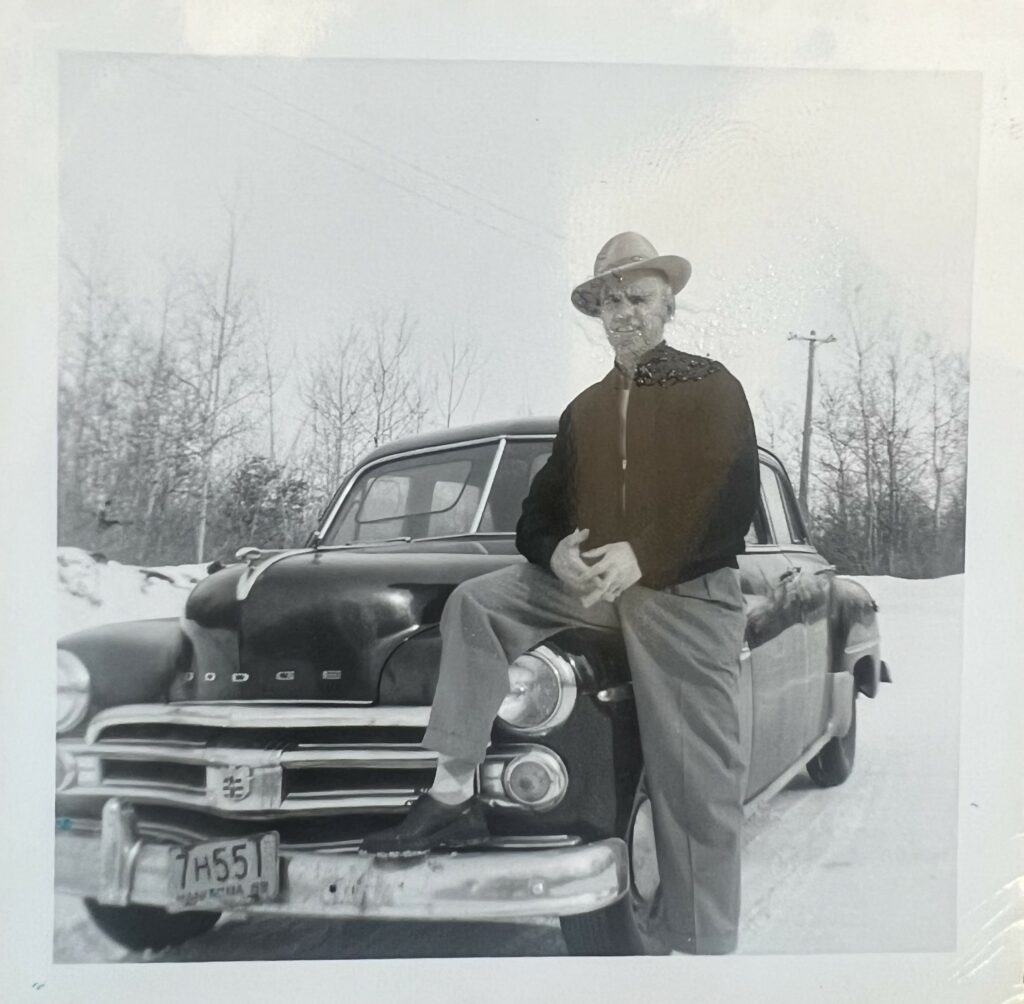 Black and white photo of a man wearing a hat standing next to an Dodge. He has one leg up on the bumper of the car and his hands clasped in front of him