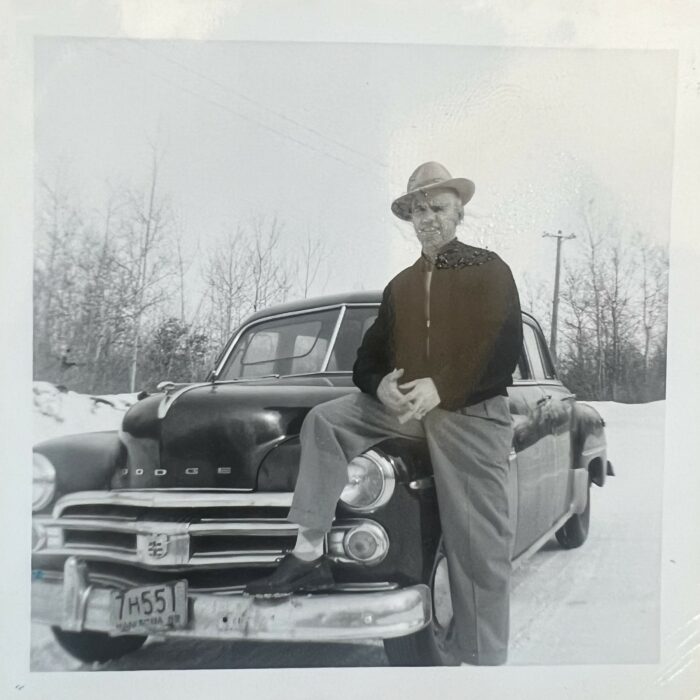 Black and white photo of a man wearing a hat standing next to an Dodge. He has one leg up on the bumper of the car and his hands clasped in front of him