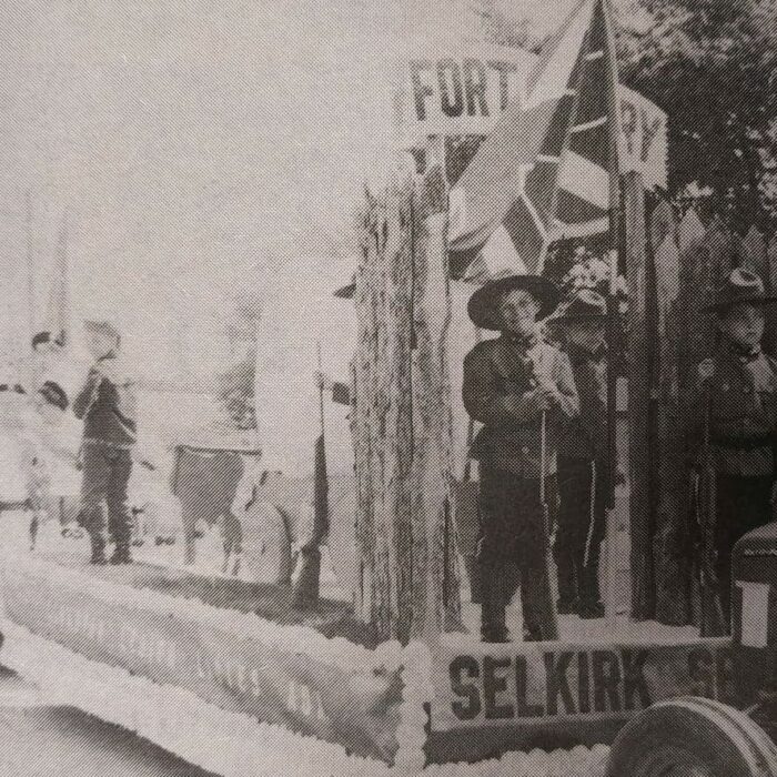 Black and white photo of a parade float. There is someone standing on the float holding a flag