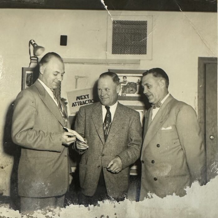 A black and white photo of three men wearing suits and ties. They all look at what the man on the left is holding