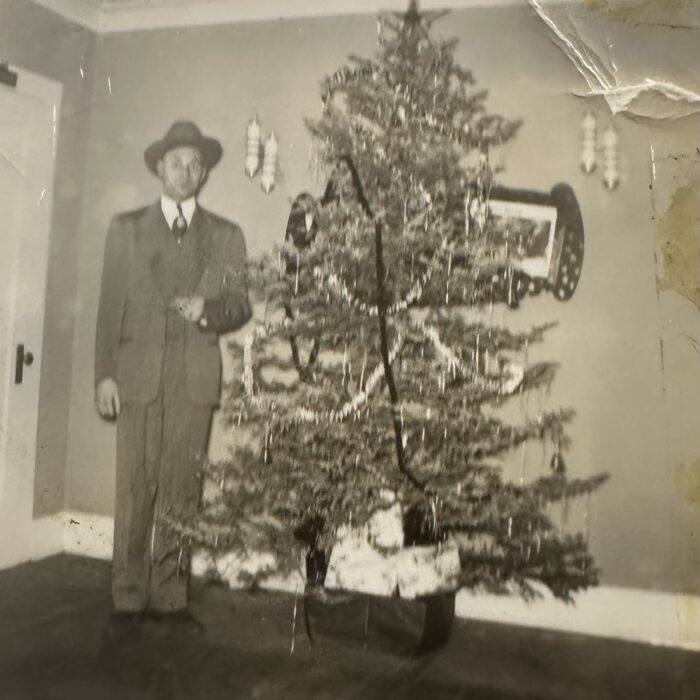 Black and white photo of a man wearing a suit and hat standing next to a decorated Christmas tree. In the background is a wall with decorations hung on the wall.