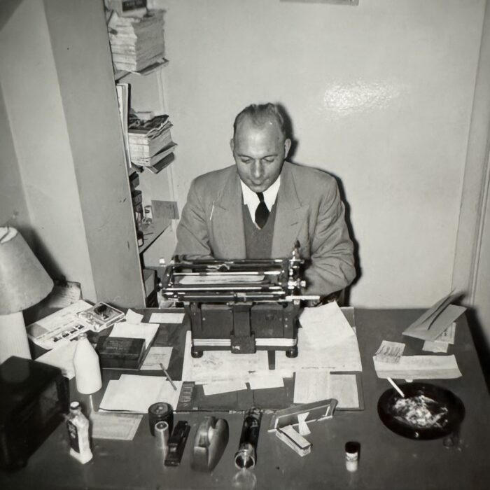A black and white photo of a man wearing a suit and tie sitting at a desk. The man types at a typewriter
