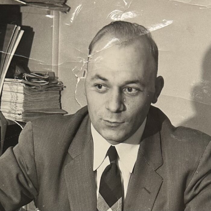 Black and white photo of a man wearing a grey suit and black tie. He is seated and looks to the left