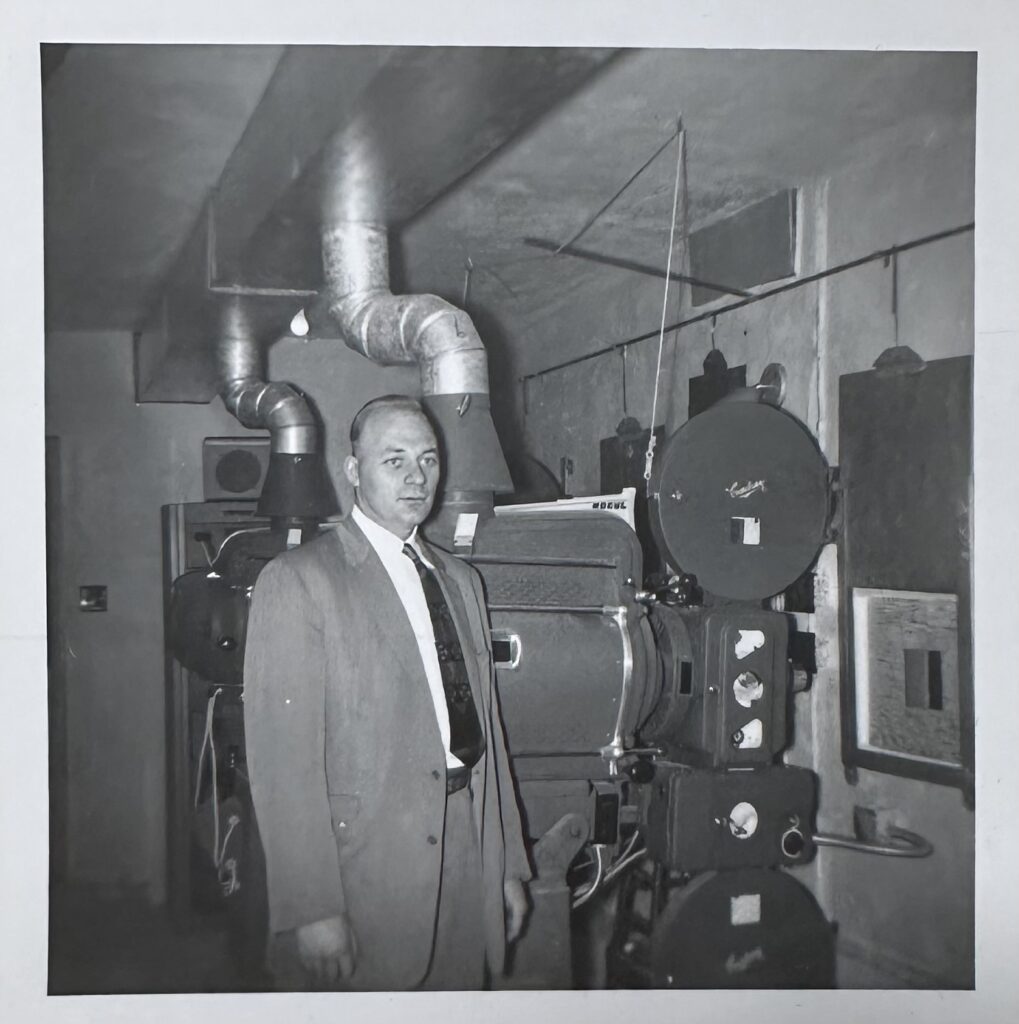 A black and white photo of a man wearing a suit and tie standing in the projection room at the Garry Theatre.