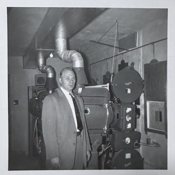 A black and white photo of a man wearing a suit and tie standing in the projection room at the Garry Theatre.