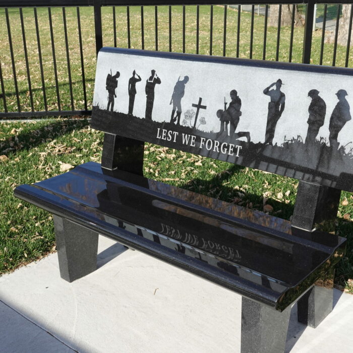 Colour photo of a black stone bench. The bench is engraved with an image of soldiers saluting and crouched around a cross