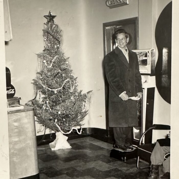 A black and white photo of a man wearing a suit and tie. He stands in the lobby next to a decorated Christmas tree