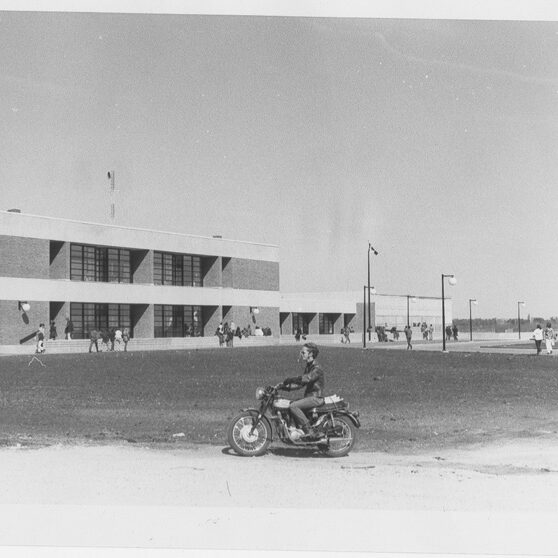 Black and white photo of a long rectangular, two-story building. There is a child on a bike at the center of the photo
