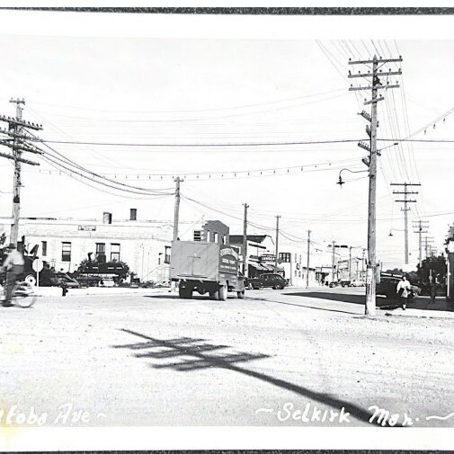 Manitoba Avenue Looking East from Main Street, 1930s, Paula Fuller Black and white photo of Manitoba Avenue. There are two large telephone poles on either side of the photo. There is a shadow of a third telephone pole diagonally across the foreground. The Selkirk Post Office is on the right hand side of the image