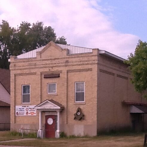 A colour image of a large brick building. The front door is red and framed by two columns which support a small pediment roof. On the building there are three windows, two on either side of the door and a third directly above the door